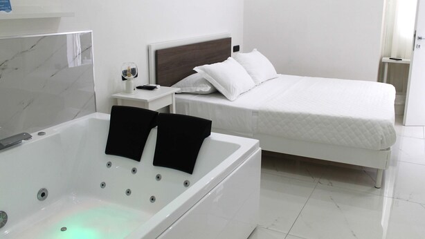 Quarto moderno com uma cama grande branca, uma mesinha de cabeceira branca, uma banheira branca e toalhas pretas.