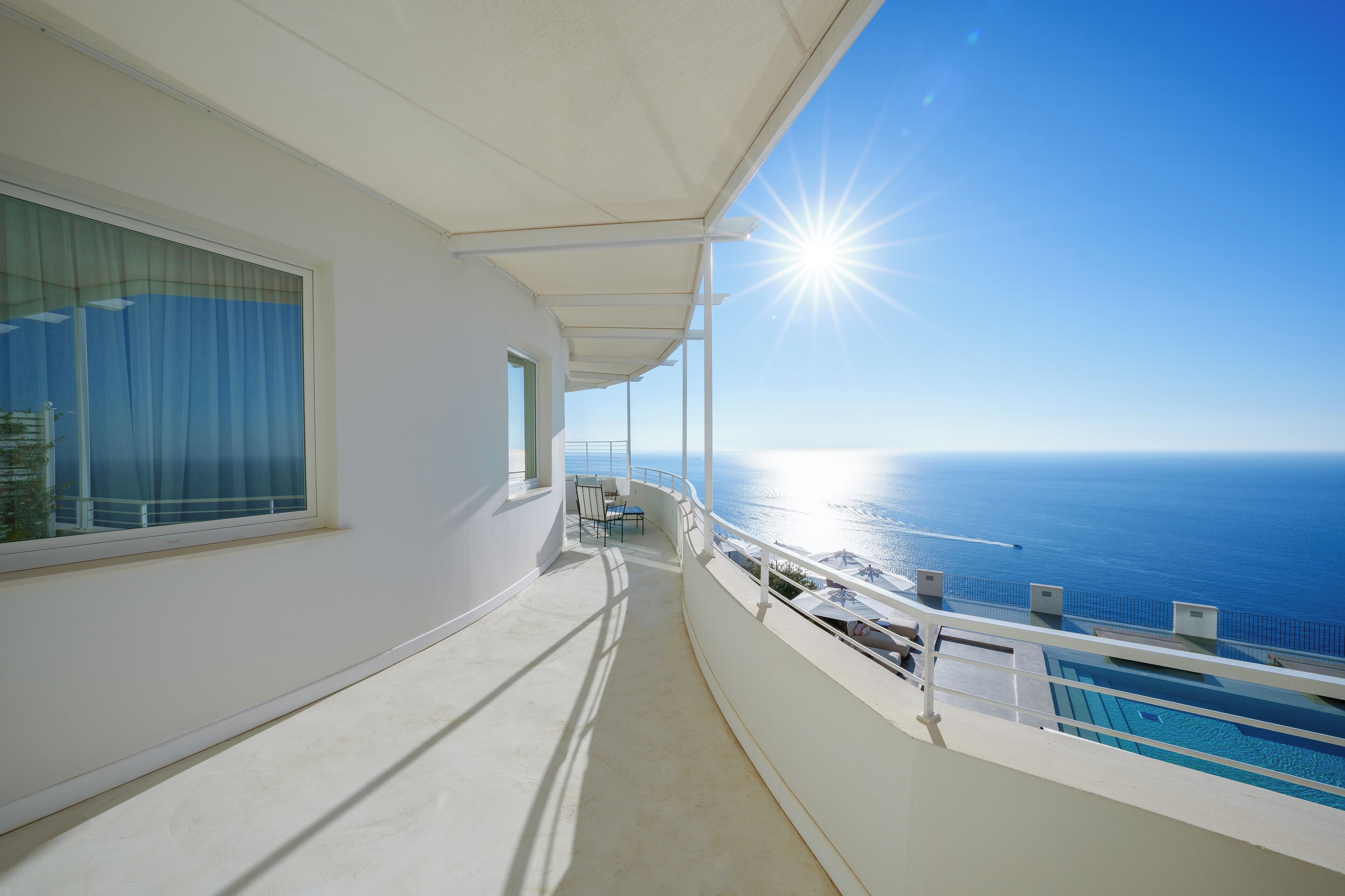 A balcony with a pool, a clear view of the sea, and a bright sun.