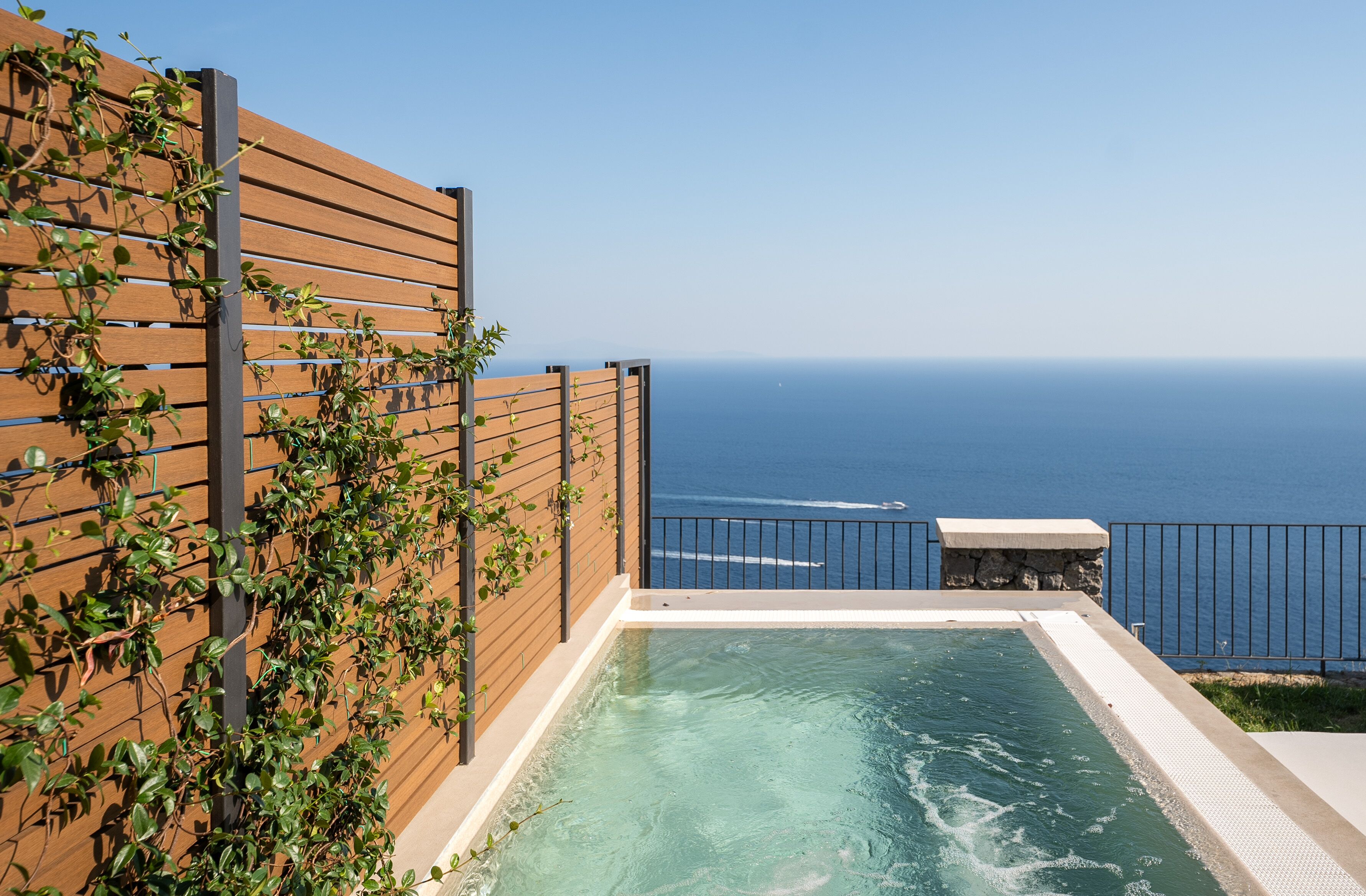 A rooftop infinity pool with a wooden deck and a stone bench, overlooking the ocean.