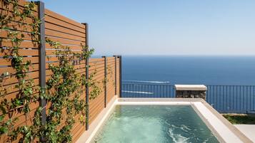 A rooftop infinity pool with a wooden deck and a stone bench, overlooking the ocean.
