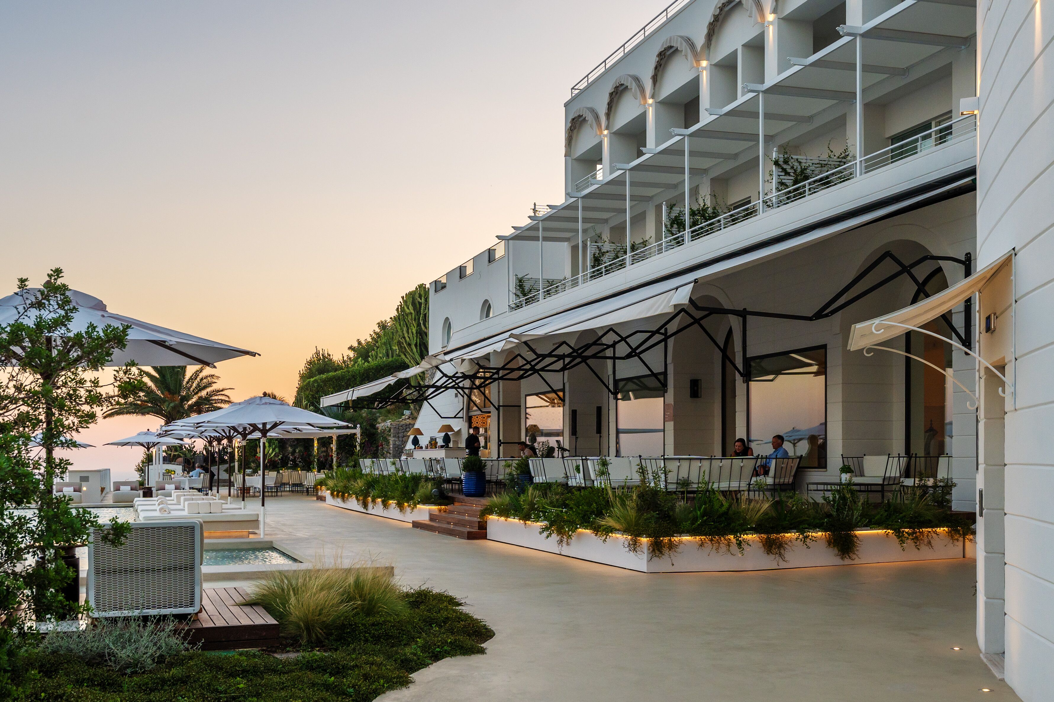 A modern outdoor dining area with multiple tables and umbrellas, surrounded by lush greenery and a pool.