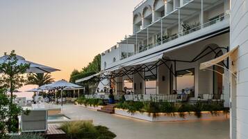 A modern outdoor dining area with multiple tables and umbrellas, surrounded by lush greenery and a pool.