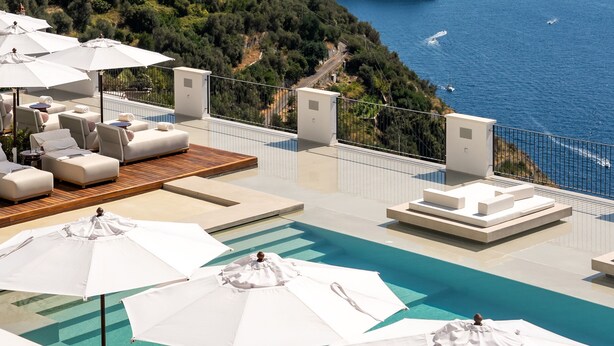 Ein Poolbereich auf dem Dach mit Liegestühlen und Sonnenschirmen, mit Blick auf eine Küstenlandschaft und eine felsige Insel im Meer.