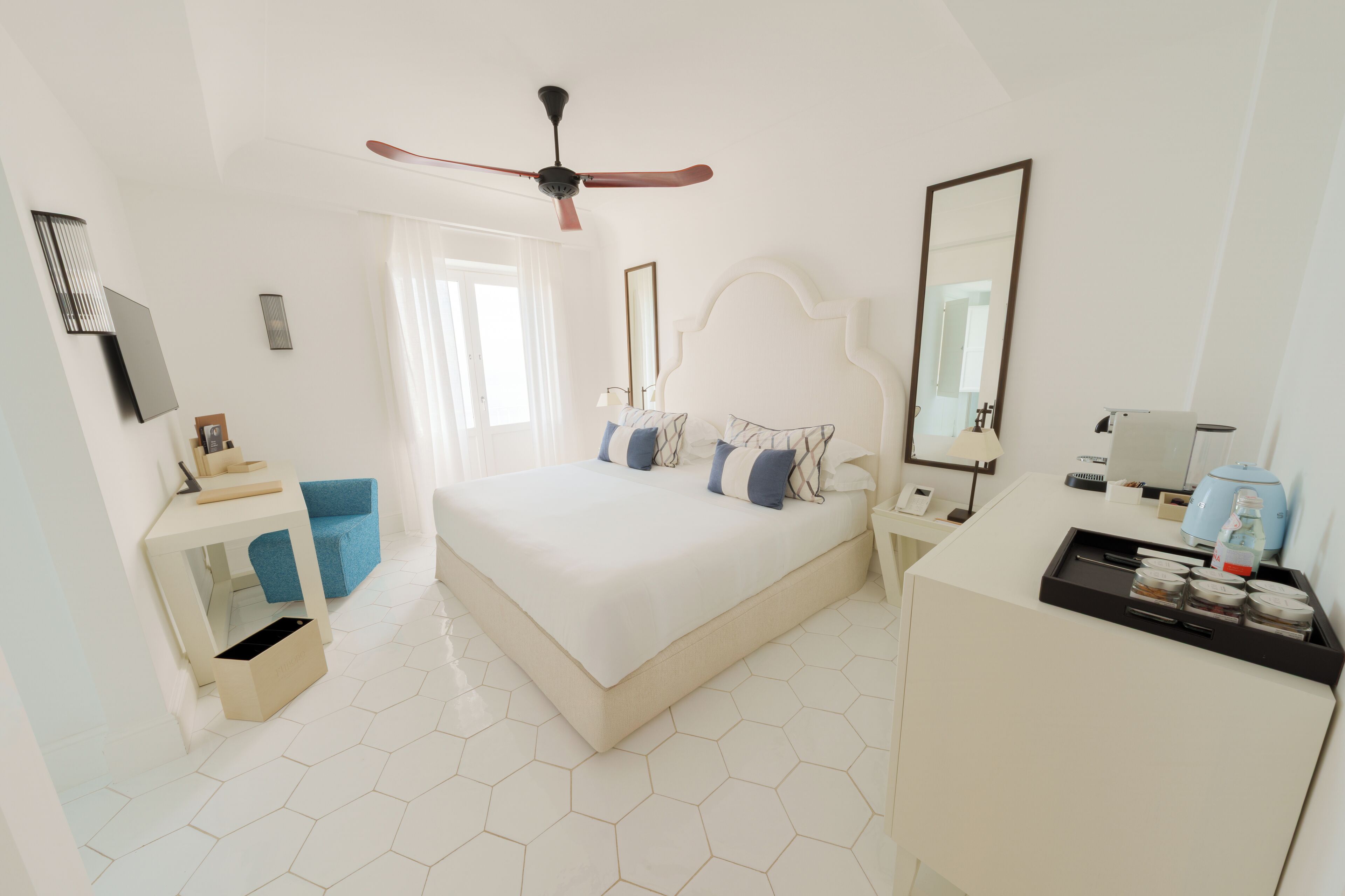 A hotel room with a large bed, a desk, a wardrobe, a ceiling fan, and a bathroom mirror.