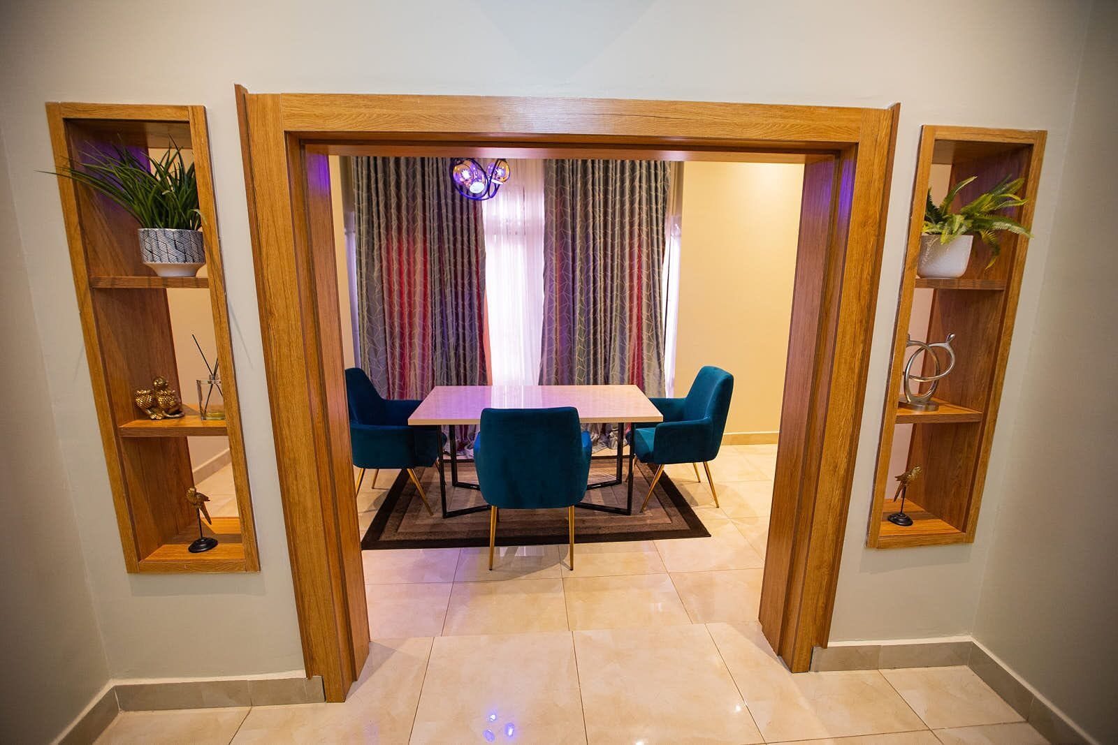 A dining area with a wooden-framed table and four blue chairs, flanked by wooden shelves with plants and decorative items.