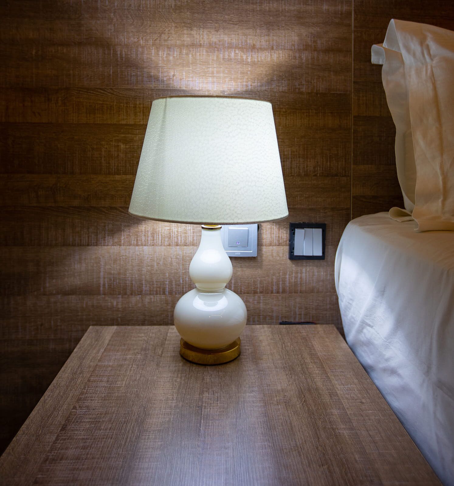 A hotel room with a wooden headboard, a bedside table with a lamp, and a bed with a white comforter.