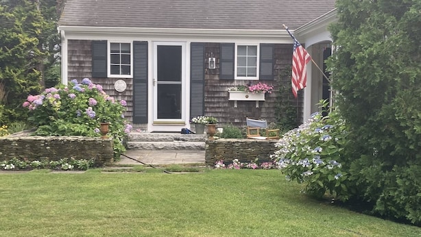 A two-story house with a dark shingle roof, a front porch with a stone wall, and a flag. -  Charming Cape Cod home, minutes to Nantucket Sound (Harwich)