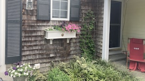 A house with dark wooden siding, a window with shutters, a planter with flowers, and a red chair outside. -  Charming Cape Cod home, minutes to Nantucket Sound (Harwich)