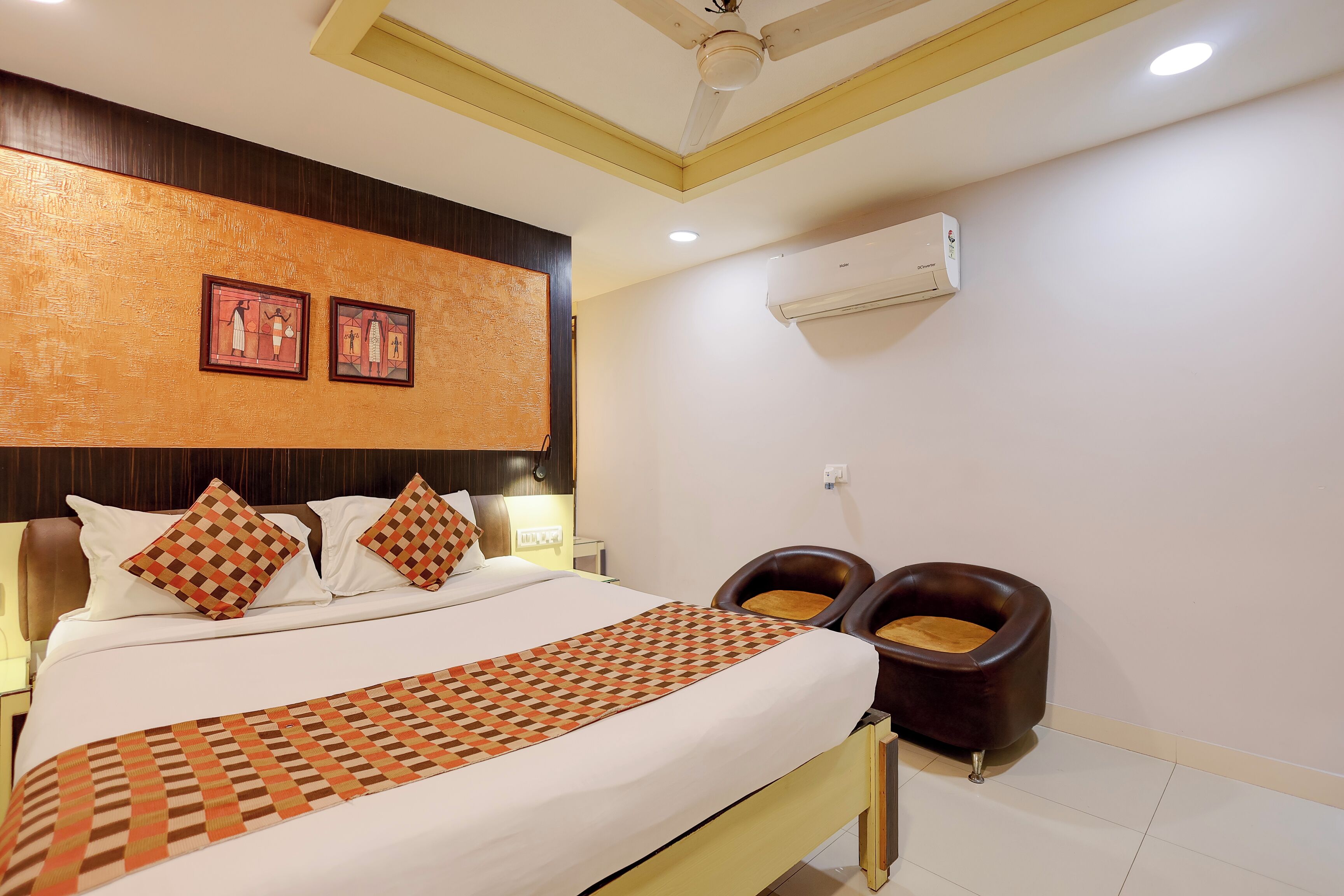 A hotel room with a bed, two wall-mounted air conditioners, a ceiling fan, and a wall with two framed pictures.