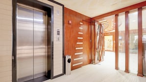 Ein moderner Aufzug mit Metalloptik, eine Holztür mit Querstreben und ein Mülleimer daneben.