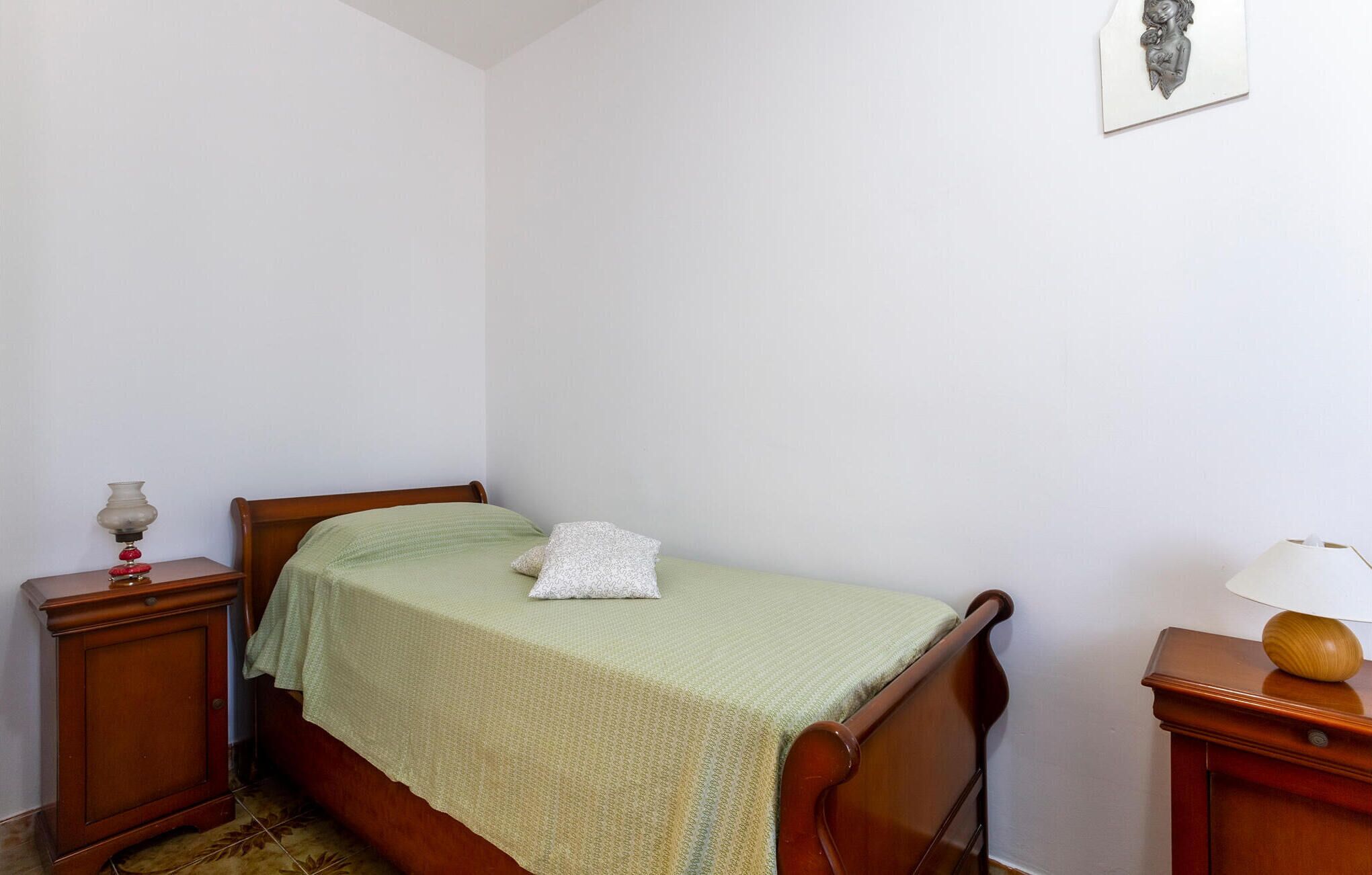 Una cama pequeña y bien hecha con una colcha verde y un almohadón blanco en una habitación sencilla con paredes blancas.