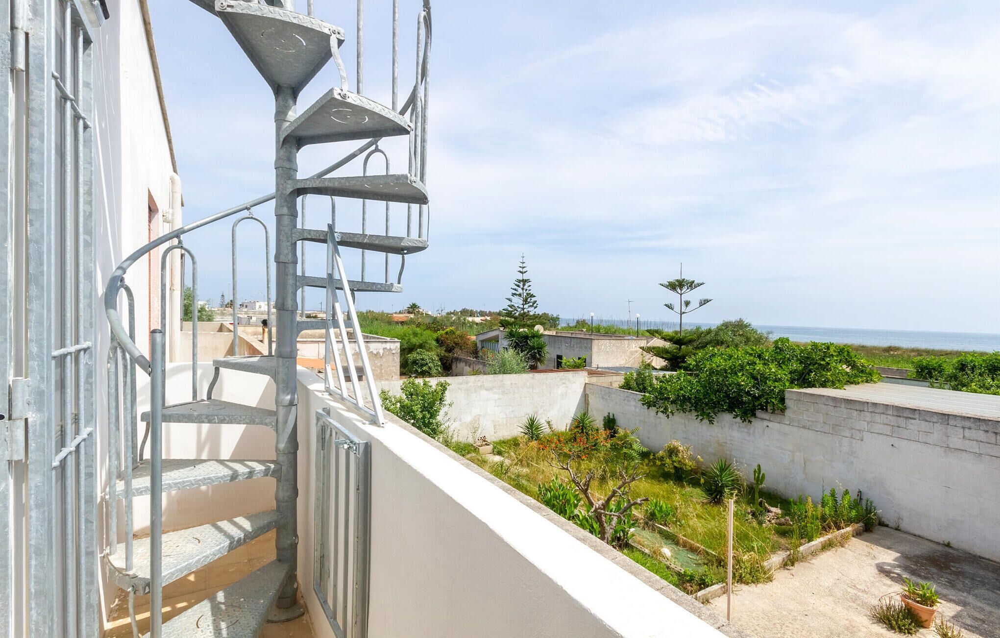 Ein Balkon mit einer Metall-Wendeltreppe, mit Blick auf eine Küstenlandschaft mit Gebäuden und Vegetation.