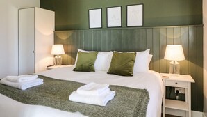 A neatly made bed with white linens and a green throw blanket, flanked by two bedside tables with lamps, in a room with a green accent wall and framed artwork above the bed.