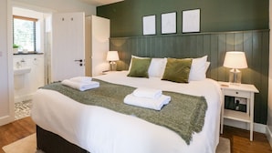 A neatly made bed with white linens and green pillows, a bedside table with a lamp, and a window with blinds.