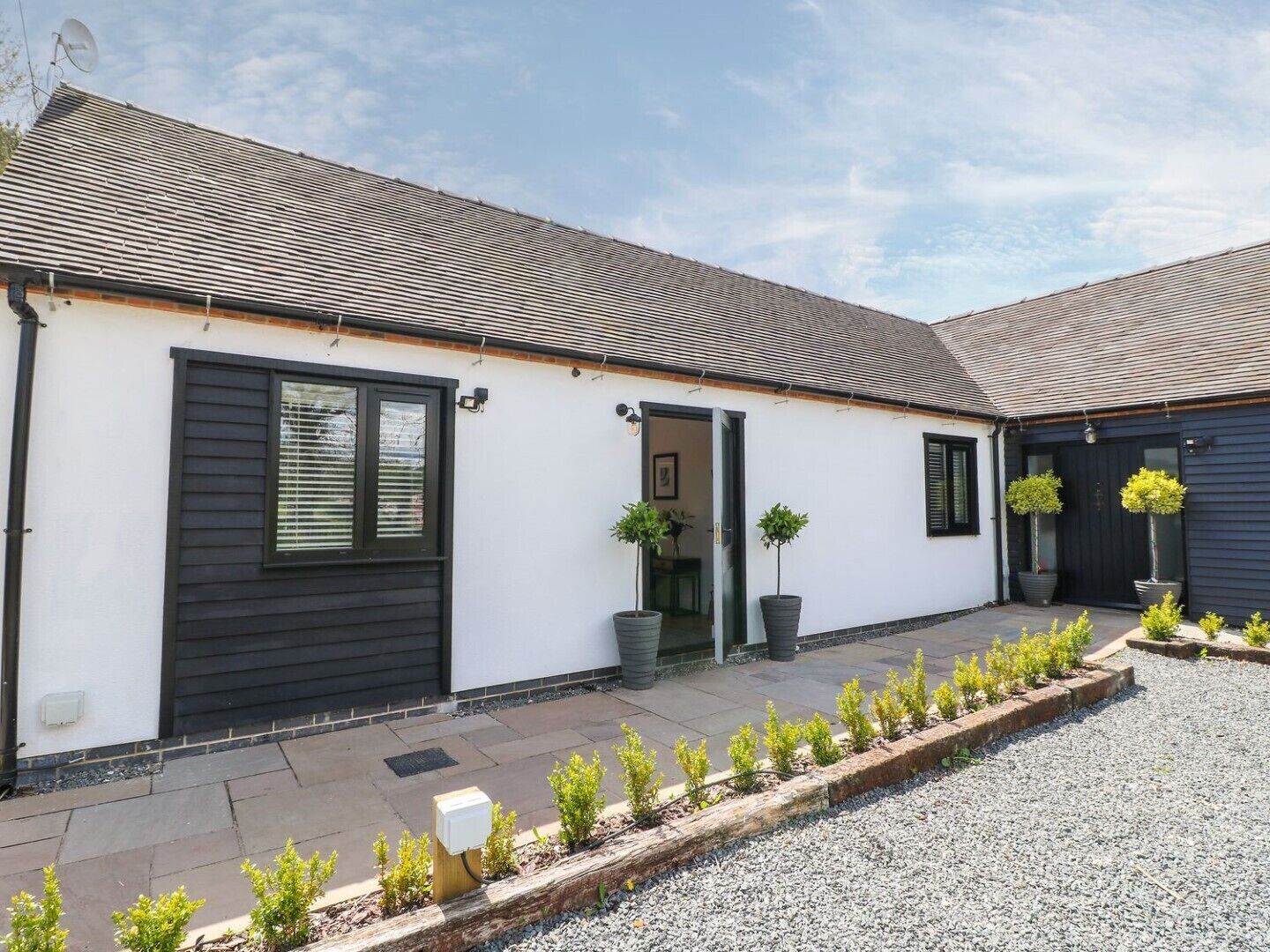 Ein eingeschossiges Haus mit einer dunkel gefärbten Haustür, einer kleinen Veranda mit Topfpflanzen und einem Schotterweg.