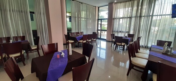 Uma sala de jantar de hotel com várias mesas e cadeiras, janelas amplas com cortinas e um interior limpo e bem iluminado.