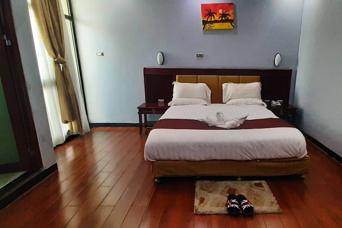 Ein Hotelzimmer mit einem großen Bett, Holzboden, einem Nachttisch, einem Beistelltisch und einem Paar Hausschuhen auf dem Boden.