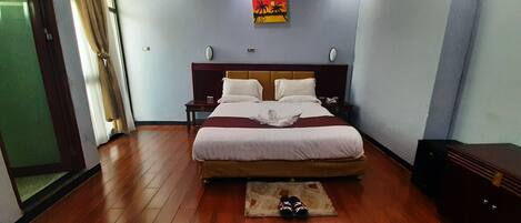 Habitación de hotel con una cama grande, suelo de madera, una mesita de noche y un par de zapatillas en el piso.