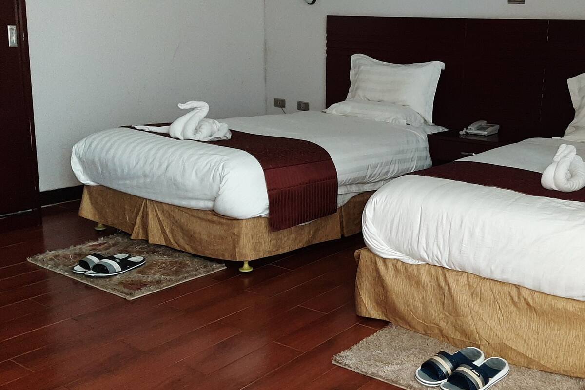Zwei Betten mit weißen Laken und braunen Betttüchern, ein Holzboden, ein Paar Hausschuhe und ein kleiner Teppich.