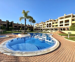 Un hôtel doté d’une grande piscine, de palmiers et d’un chemin bordé de briques.