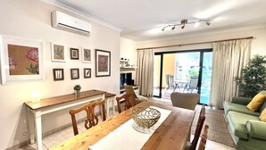 Uma área de refeições com uma mesa de madeira e cadeiras, uma mesinha lateral com um abajur, e um sofá verde ao fundo.