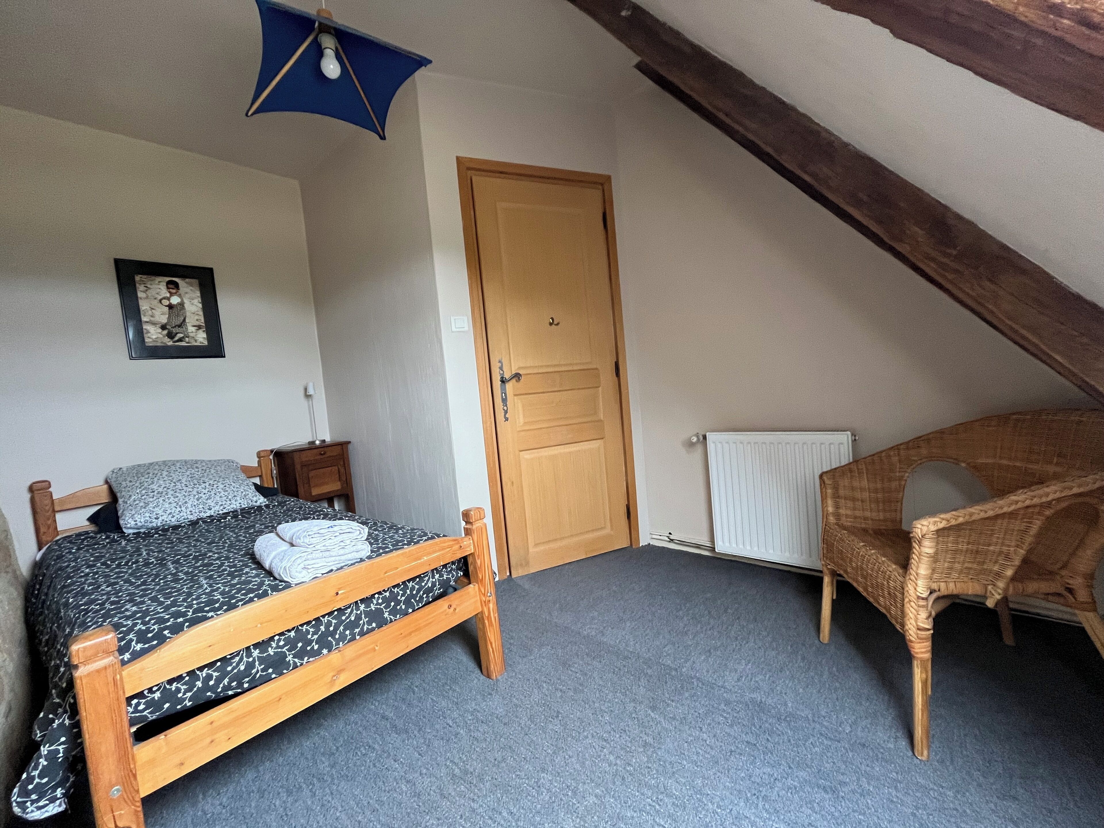 Un dormitorio con una cama, una puerta de madera, una silla de mimbre y un cuadro enmarcado en la pared.