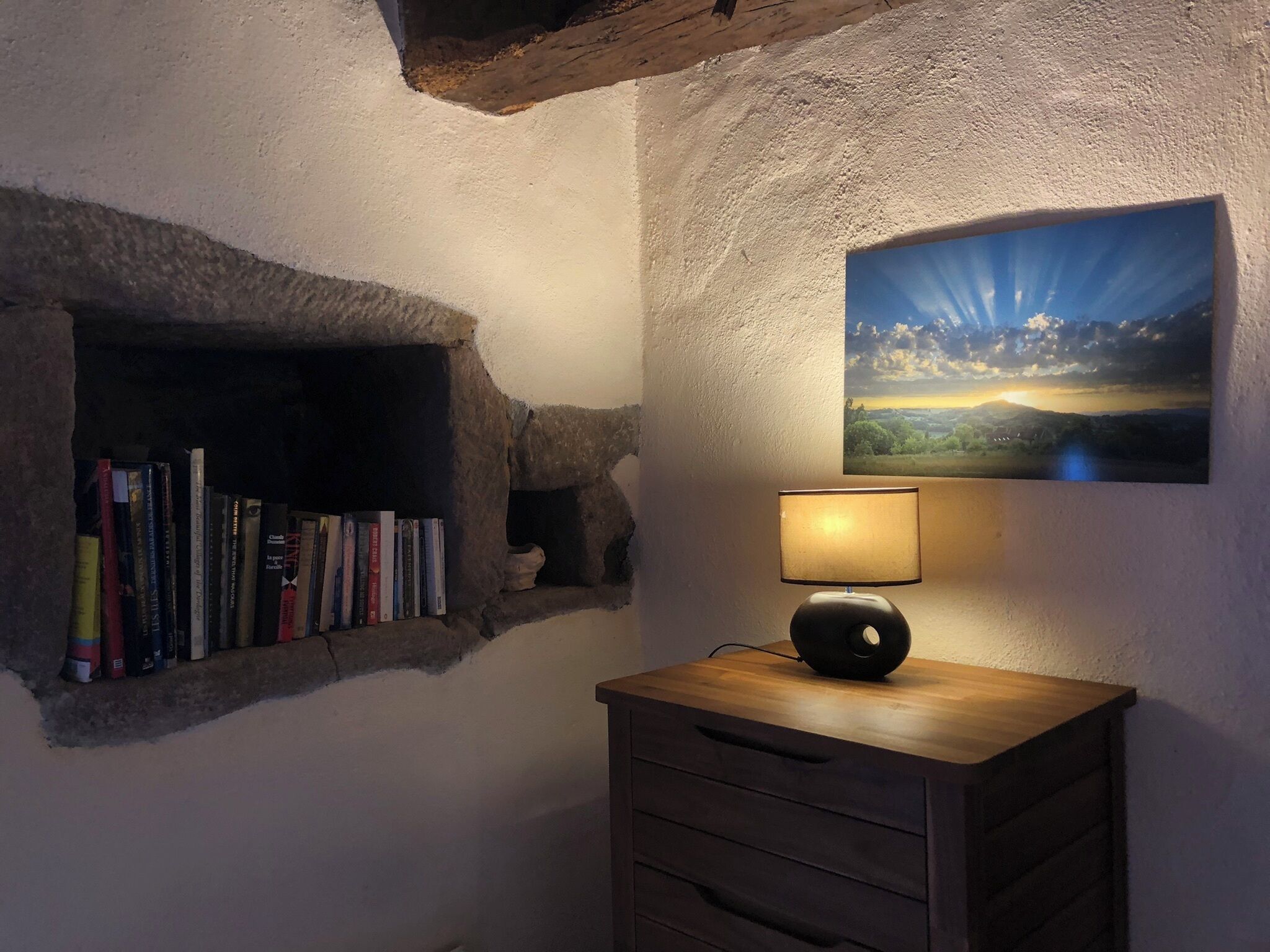 A cozy room with a stone shelf holding books, a wooden cabinet with a lamp, and a landscape painting on the wall.