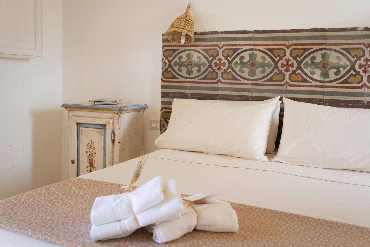A neatly made bed with white linens and a patterned bedspread, a small bedside table with a book, and a decorative wall panel.