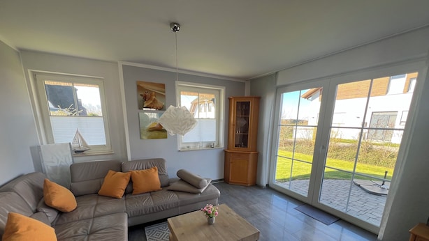 Un salon moderne avec un canapé d’angle gris, une table basse en bois et une grande porte vitrée donnant sur une terrasse extérieure.