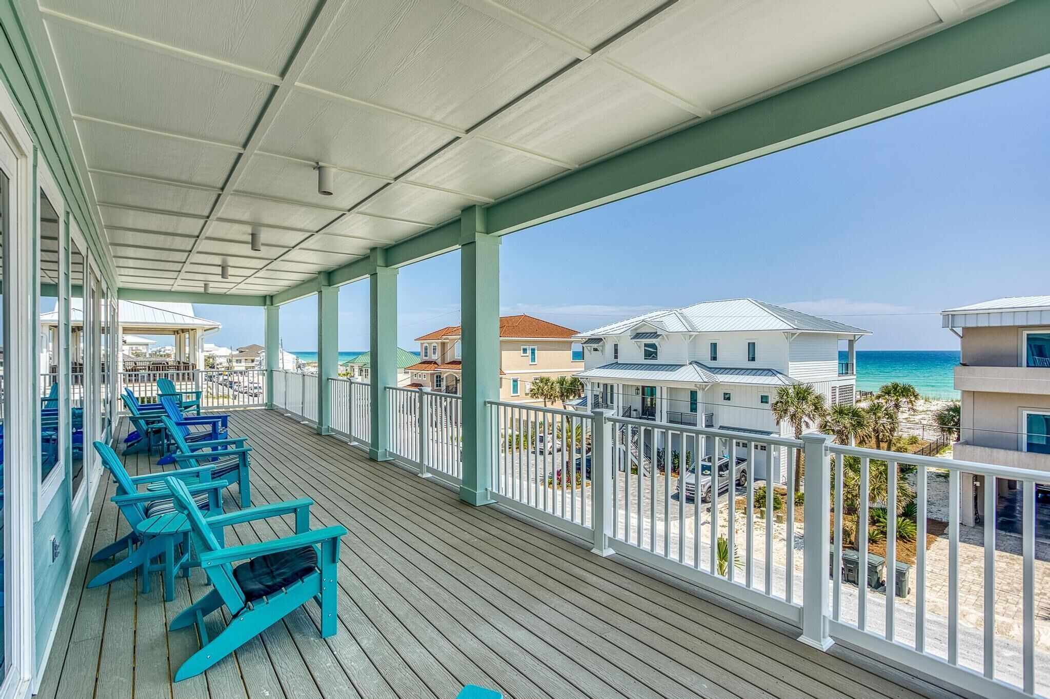 A covered balcony with blue chairs, a view of the beach, and houses in the background.