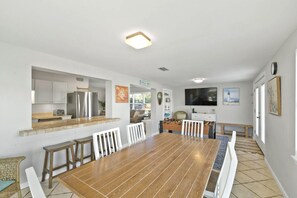 Un vaste espace de salle à manger avec une longue table en bois et des chaises blanches, une cuisine équipée d’appareils modernes et un coin salon confortable avec un canapé et une table basse.