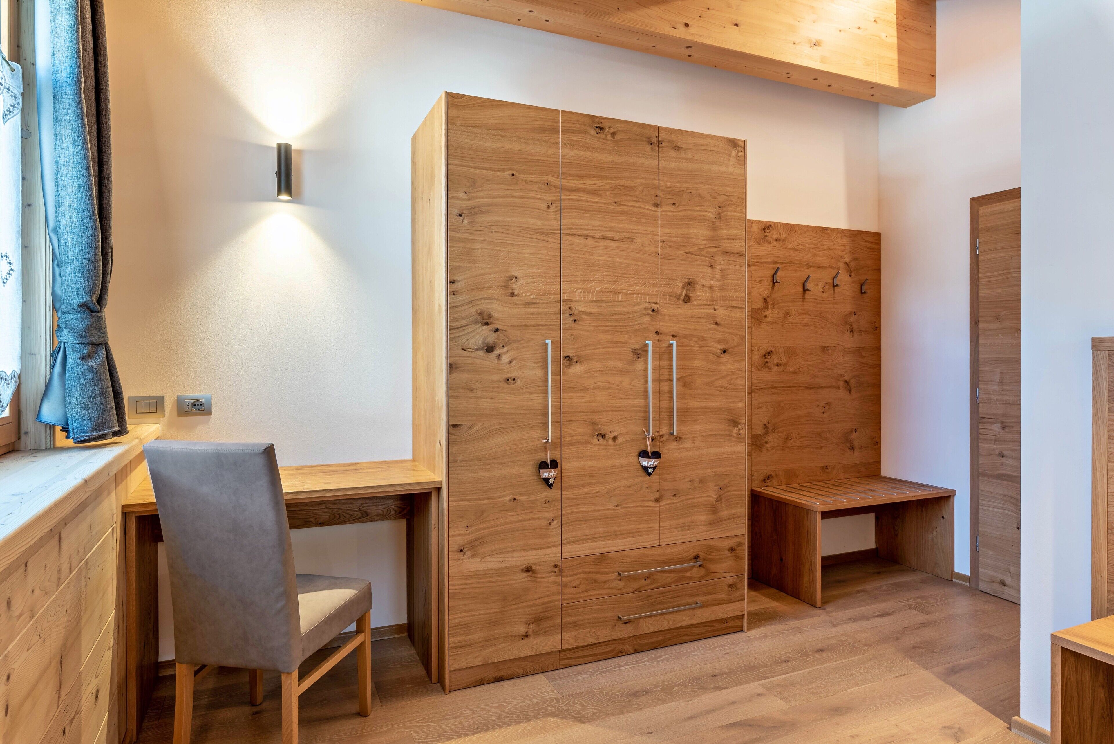 A room with a wooden wardrobe, a desk, a chair, and a bench.