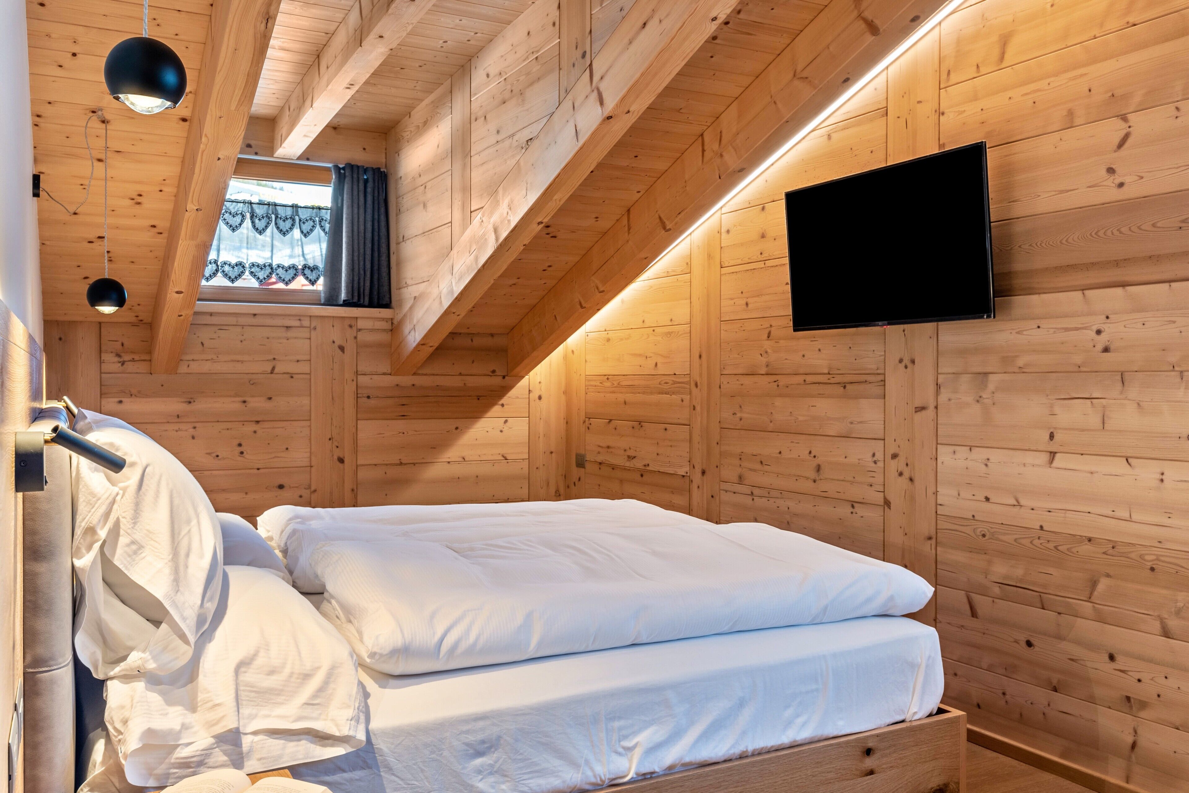 A wooden cabin room with a bed, a television mounted on the wall, and a window with curtains.