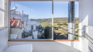Une pièce dotée d’une grande fenêtre donnant sur un bâtiment aux toits recouverts de tuiles rouges et sur un paysage vallonné.