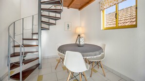 Un escalier en colimaçon, une table ronde entourée de chaises, une lampe et une fenêtre donnant sur un toit recouvert de tuiles.