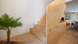 Un escalier en bois menant à un deuxième étage, une plante en pot et un coin cuisine avec des meubles blancs.