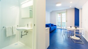 A modern bathroom with a glass shower enclosure, a white sink, and a mirror. There is a blue sofa and a round table with chairs in the adjacent living area. - Piazza Municipio Blue Apartment by Wonderful Italy (Naples)