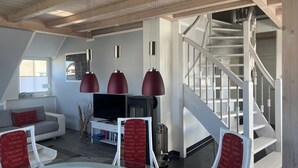Een moderne woonkamer met een witte bank, rode stoelen, een glazen salontafel en een televisie. Een trap met witte leuningen leidt naar een hoger gelegen verdieping.
