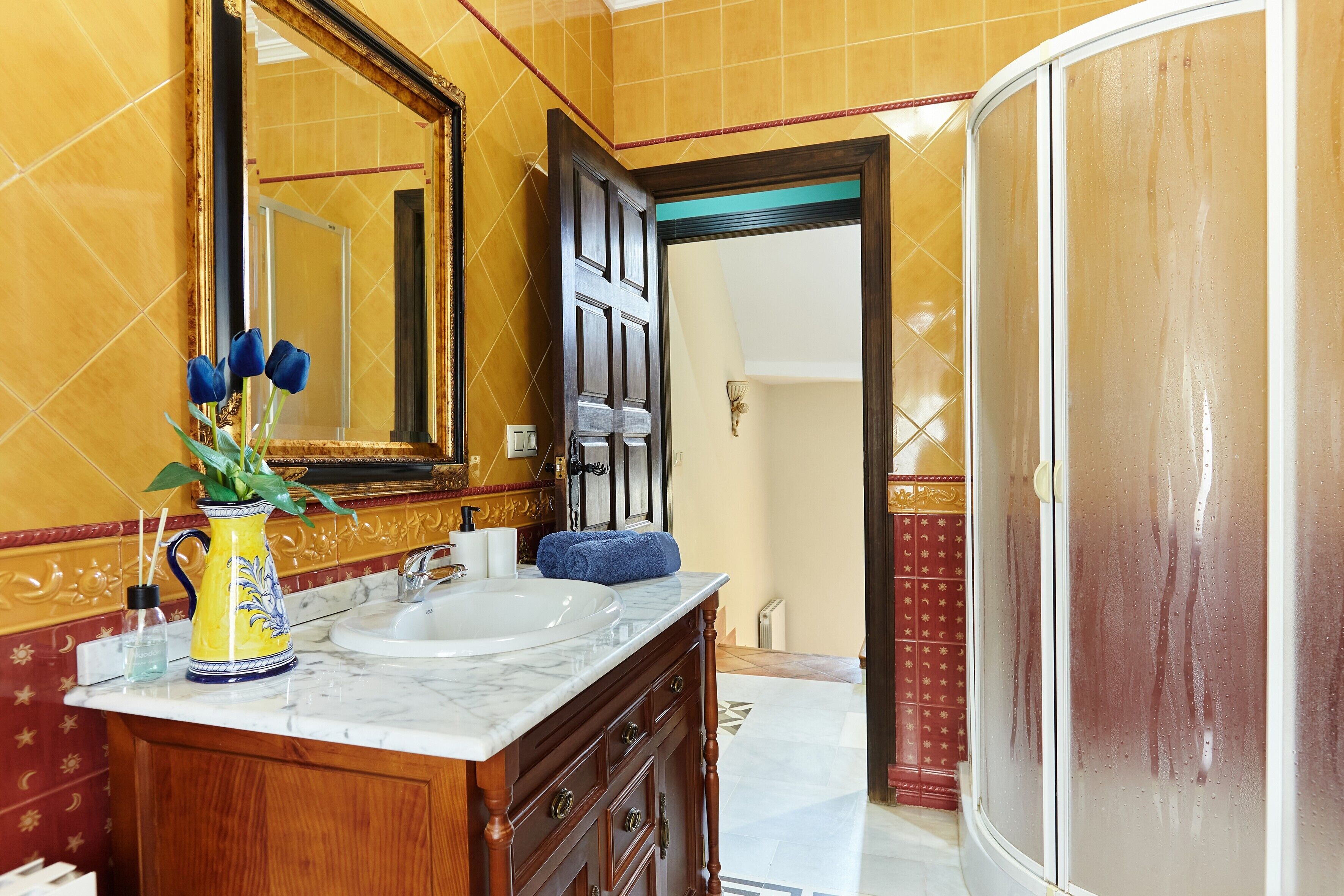 Casa de banho com duas pias, um espelho, um vaso amarelo com flores azuis e zona de duche.