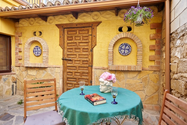 Una mesa con mantel verde, dos sillas, un libro, dos copas de vino, un jarrón con flores y una puerta de madera con detalles decorativos.