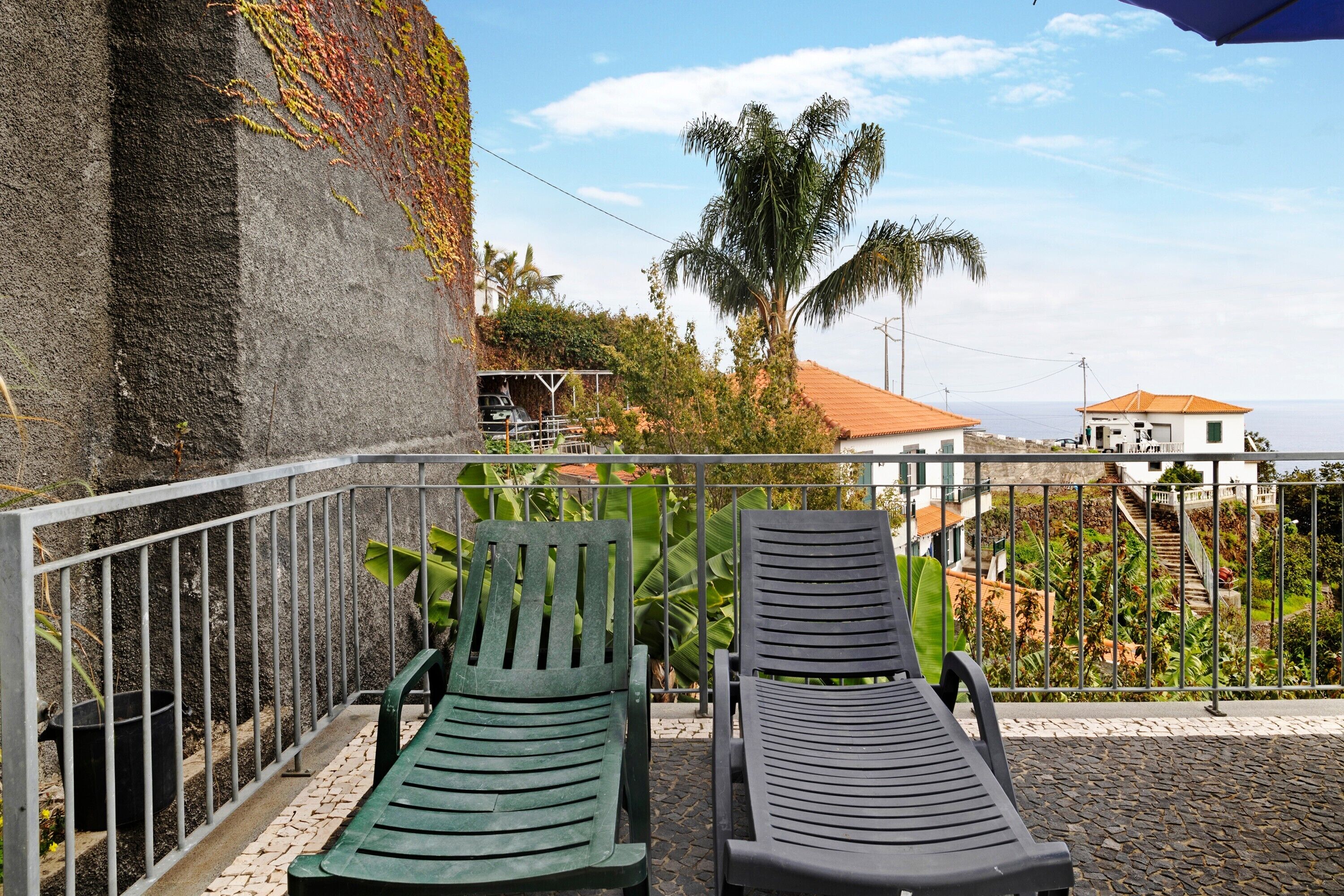 Two lounge chairs on a balcony with a view of a coastal area, including a palm tree and buildings.