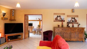Una sala de estar con televisor, un sofá rojo y amarillo, un mueble de madera y un comedor al fondo.