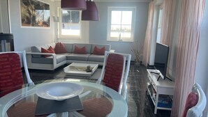 Un salon avec un canapé, une table basse en verre et un téléviseur. Il y a deux fenêtres avec des rideaux, une cheminée et une bibliothèque.