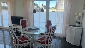 Un coin repas avec une table en verre entourée de chaises en osier, un téléviseur et un meuble sur lequel est posée une plante en pot.