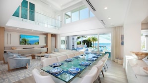 A spacious dining area with a long table set for six, a large flat-screen TV mounted on the wall, and a balcony with a view of the ocean. - Sun Rays by Grand Cayman Villas (Cayman Kai)
