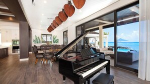 A modern living room with a grand piano, a dining area, and a large window overlooking a pool and ocean. - Rip Kai by Grand Cayman Villas (Rum Point)