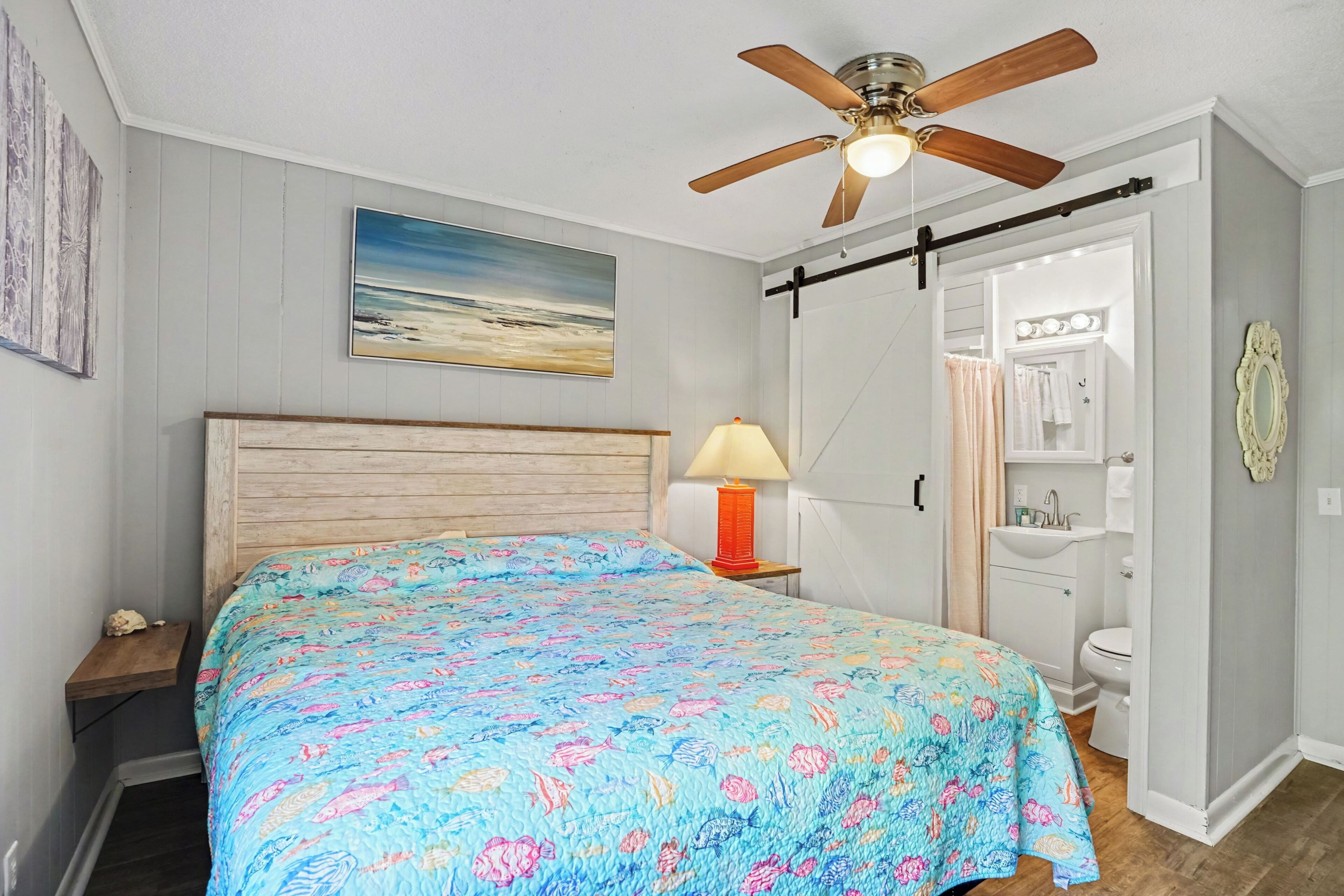 A bedroom with a bed, a ceiling fan, a nightstand with a lamp, a painting, and a bathroom with a sink and mirror.