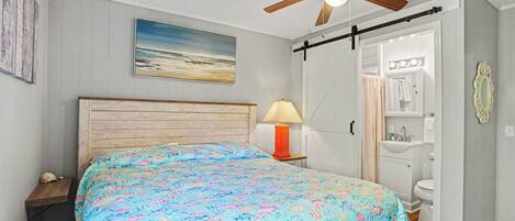 A bedroom with a bed, a ceiling fan, a nightstand with a lamp, a painting, and a bathroom with a sink and mirror.