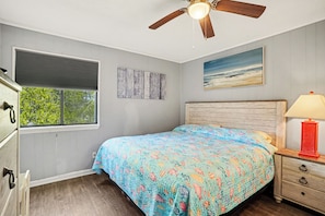 Quarto com cama, mesa de cabeceira, abajur, ventoinha no teto, janela com persianas e um quadro na parede.