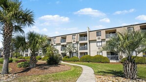 A multi-story residential building with a landscaped garden, a curved pathway, and a clear sky. - A Place At The Beach #3C Short Walk to the Beach! (North Myrtle Beach)